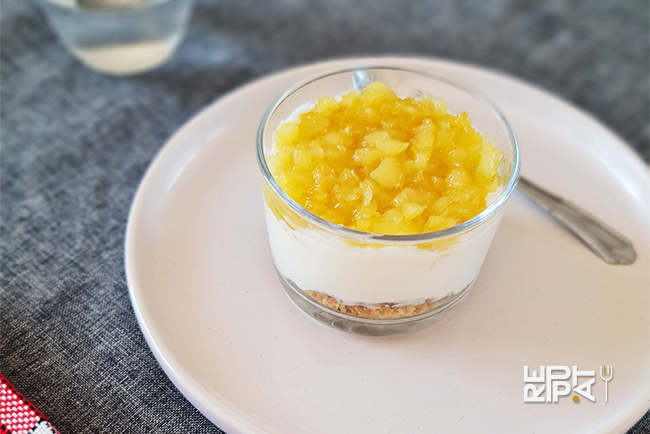 Verrine avec du biscuit écrasé, du fromage blanc et des dés d'ananas, servie dans un contenant en verre sur une assiette blanche